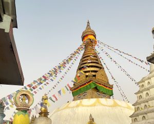 A low-angle shot of a golden Buddhist stupa with traditional wisdom eyes painted on its base. Strings of colorful prayer flags stretch outward from the tiered top spire against a clear, light sky. Smaller white dome structures and ornate stone carvings border the edges of the frame. 