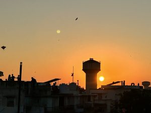 Kites fill an orange sunset sky over silhouetted rooftops, with people flying them during a kite festival.
