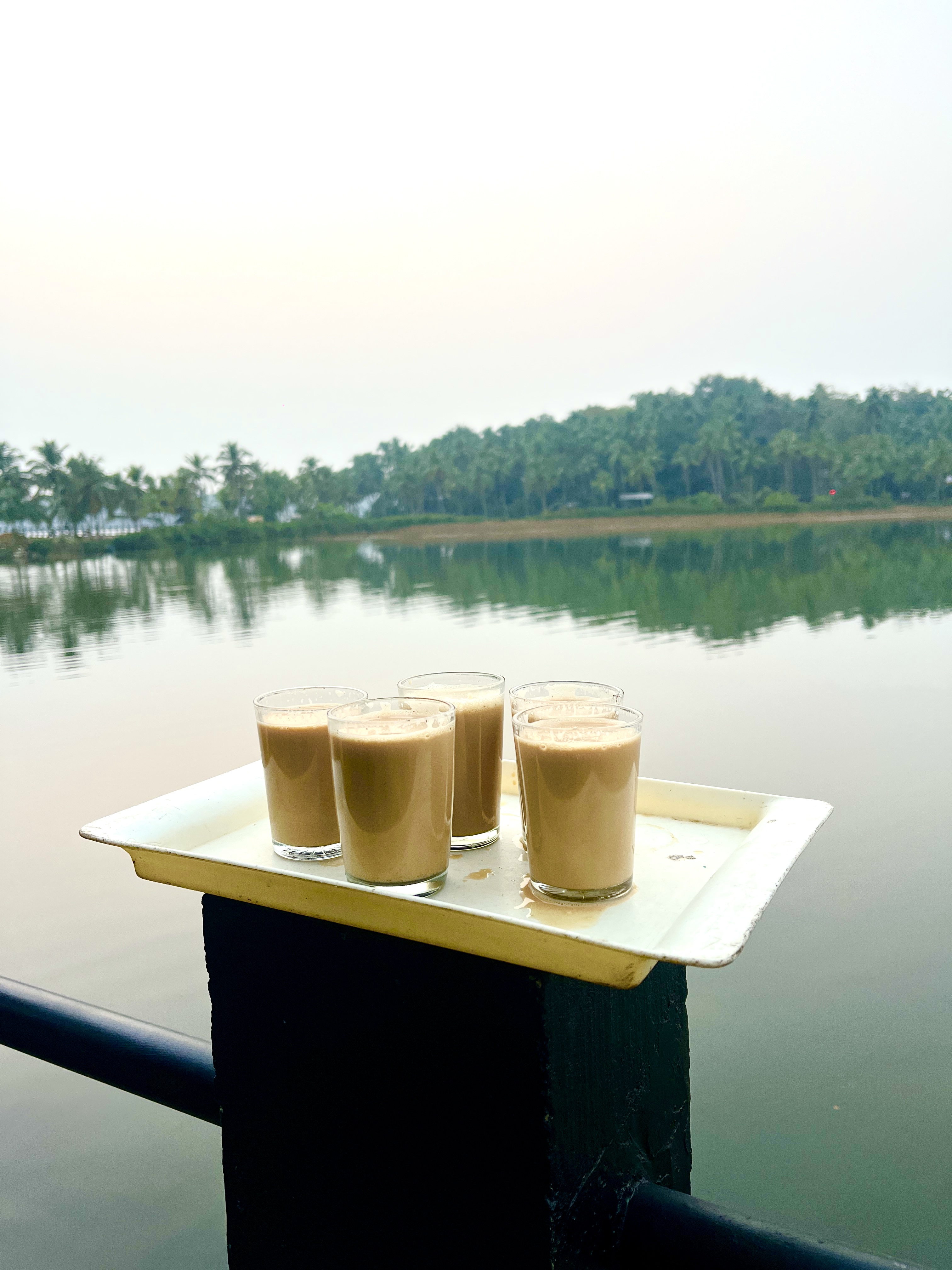 5 glasses of chai on a yellow tray sit on a railing by a serene river, with reflections of lush green trees in the calm water