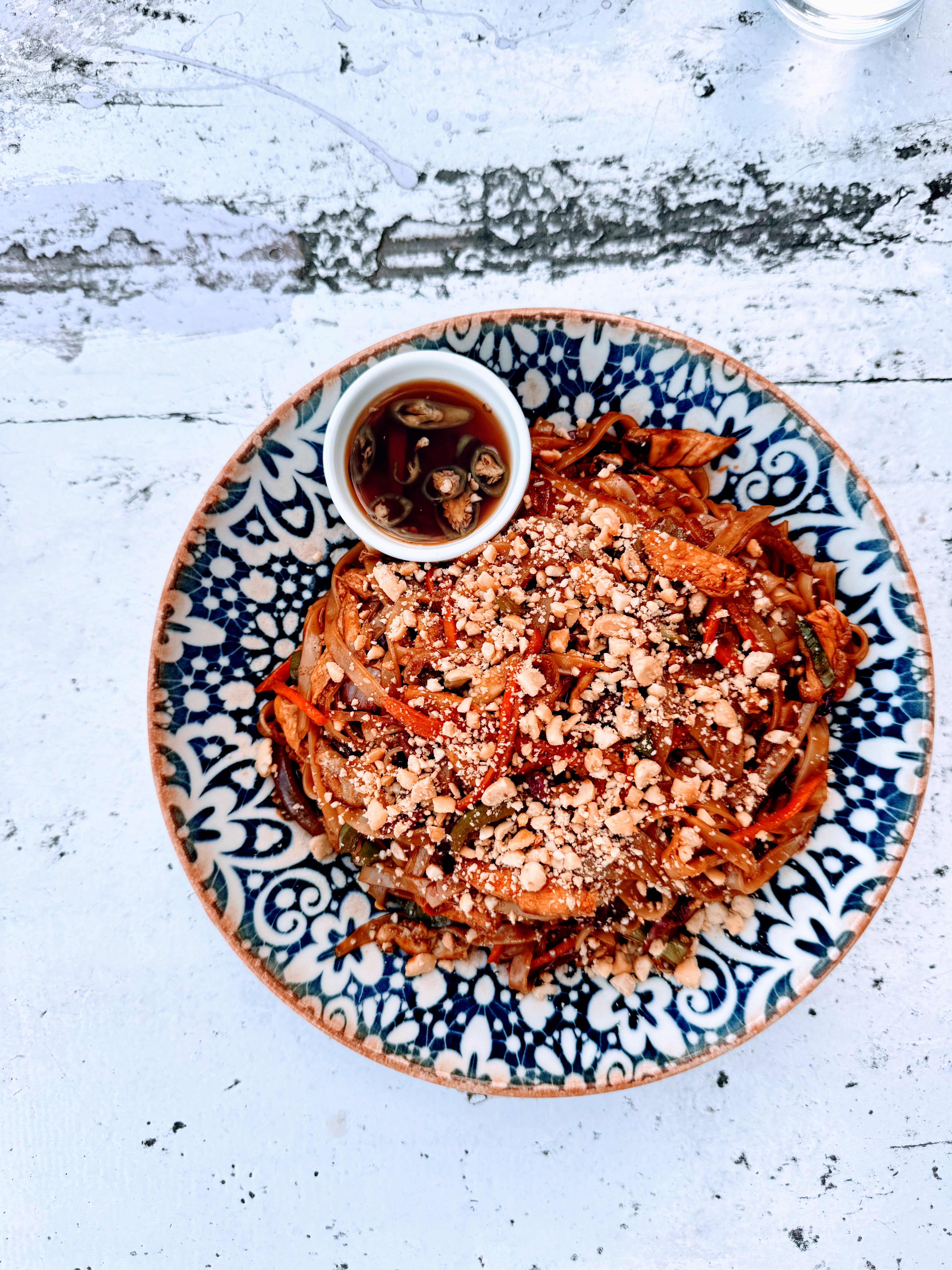 A top-down view of a plate of Pad Thai noodles topped with crushed peanuts, served alongside a small bowl of chili soy sauce on a white rustic table.