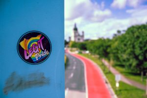 A “Legit Pride” rainbow sticker on a blue post, with a blurred road, red bike lane, trees, and distant building in the background. #shareyourpride2026
