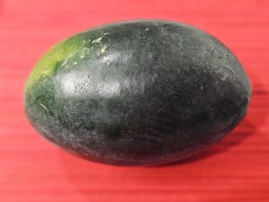A dark green watermelon is placed against a red background, creating a bold visual contrast. The smooth texture and natural shape make the fruit the clear focus of the image. Captured in Perumanna, Kozhikode, Kerala. 