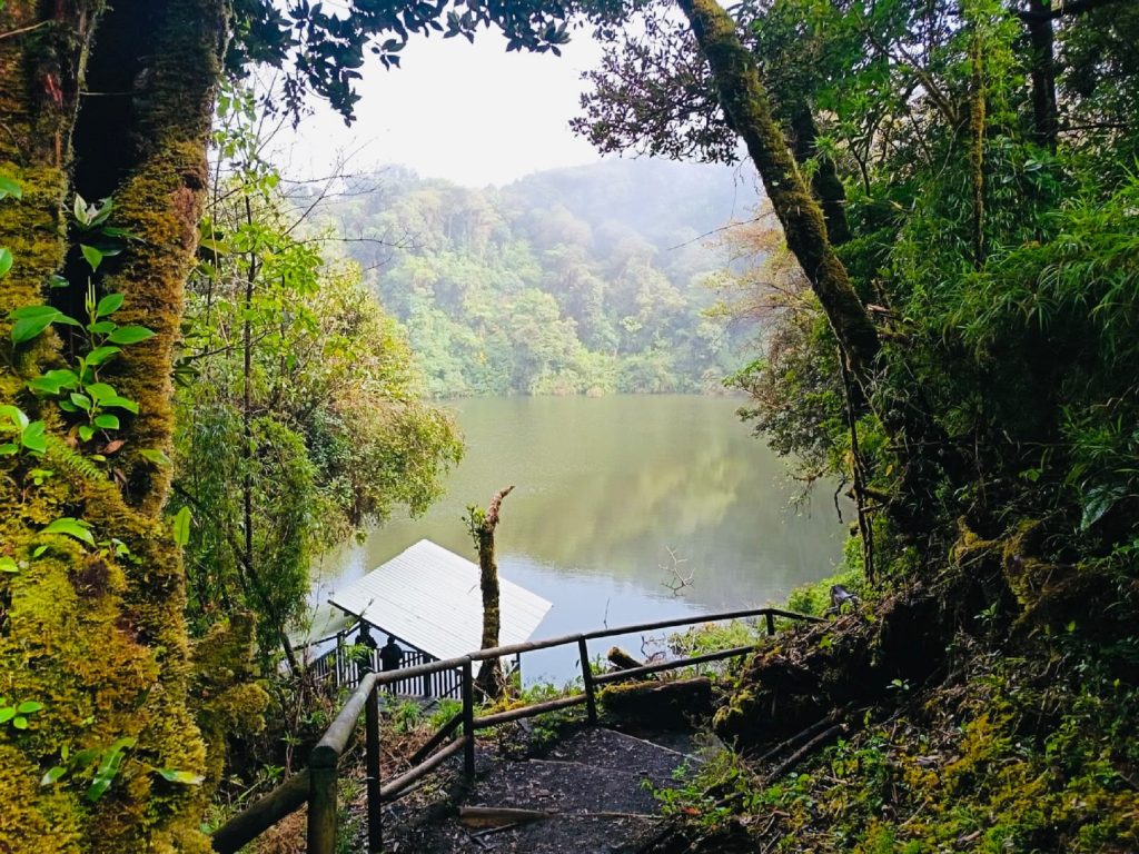 Lake Barva is surrounded by a cloud forest with moss-covered trees and abundant vegetation. The still water reflects the surrounding landscape, while a light mist hangs over the distant hills. The scene highlights the tranquility and natural beauty of this protected area.