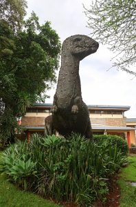A tall, textured concrete statue of a long-necked dinosaur standing amidst a bed of green snake plants.
