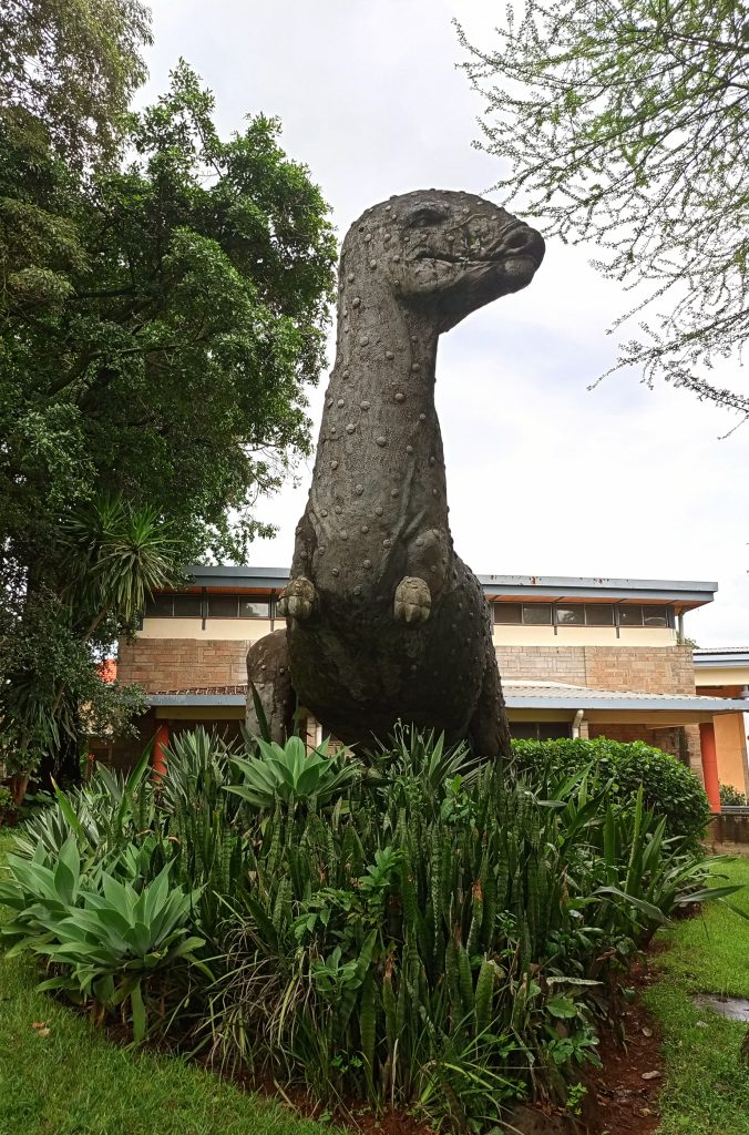 A tall, textured concrete statue of a long-necked dinosaur standing amidst a bed of green snake plants.