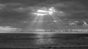 Black-and-white beach scene with dark clouds over the ocean. Sunlight breaks through a gap in the clouds, sending bright rays down onto the water. Waves roll toward the shore, with a few small figures visible in the sea.