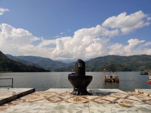Shiva linga on a lakeside temple platform overlooking Begnas Lake with forested hills and billowing clouds.