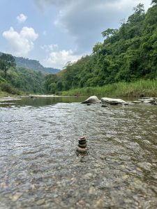 Calm Sangu river scene with rocky stack in water, surrounded by lush green forests and distant hills of Bandarban under a cloudy sky, conveying tranquility.