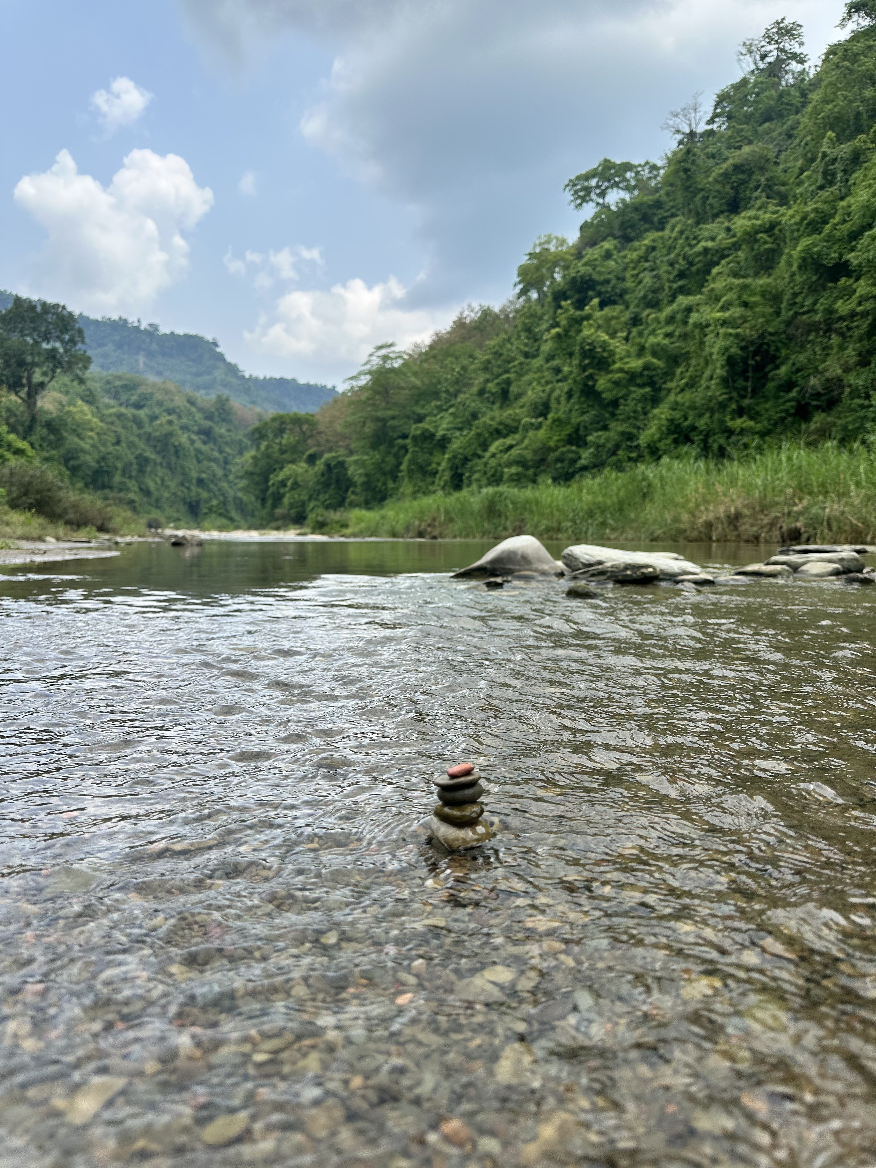 Calm Sangu river scene with rocky stack in water, surrounded by lush green forests and distant hills of Bandarban under a cloudy sky, conveying tranquility.