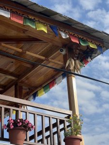 Rustic wooden balcony with colorful prayer flags and drying corn. Pink flowers in pots, under a clear blue sky, create a peaceful, cultural scene.
