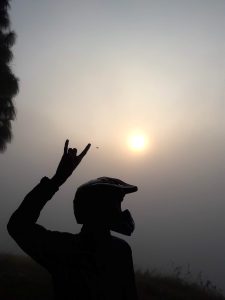 Silhouette of a person in a motorcycle helmet making a sign with their hand raised against a hazy morning sun in a misty sky. 