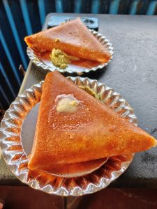 Two crispy triangular butter masala dosas served on disposable plates, topped with melting butter and accompanied by green chutney. The photo captures a simple street food moment with warm colors and a casual local dining feel.
