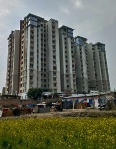 High-rise apartment buildings beside a yellow mustard field under a cloudy sky in an urban residential area.
