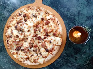 A whole pizza topped with melted cheese, sliced mushrooms, sausage pieces, and shredded meat sits on a round wooden peel. To the right of the pizza, a small metal bucket contains a burning orange candle with a small sprig of rosemary tucked into the handle. The items rest on a dark green marbled surface. 
