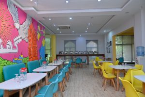 Bright café interior with colorful decor. Vibrant pink mural with a bird on the left, turquoise and yellow chairs, white tables, and buffet in the back

