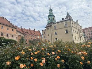A charming historic courtyard framed by elegant old buildings, with vibrant orange roses in bloom under a softly clouded sky.
