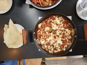A round black metal skillet featuring oven Kofte meatballs cooked slowly with a vibrant tomato-based vegetable mixture and topped with melted cheese served alongside a plate of folded flatbread.