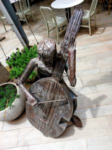 A stylized bass player figure sculpture crafted entirely from welded scrap metal, depicted in a seated posture while playing a double bass with a metal bow. 