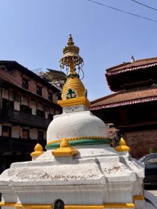 A white Buddhist chorten (stupa) with the all-seeing eyes of Buddha, golden spire, and green and yellow decorative bands, set against a clear blue sky with traditional Nepali pagoda rooftops in the background. 