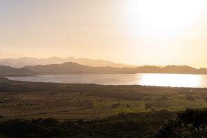 A wide landscape view of a lake surrounded by rolling mountains and open fields. Golden sunlight reflects brightly on the water, with layered hills fading into the hazy sky in the background.
