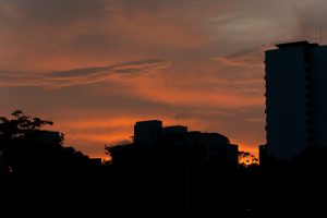 An orange sunset sky with soft clouds glowing above dark silhouettes of buildings and trees. A tall building stands on the right side, while the city skyline appears mostly in shadow against the bright horizon.