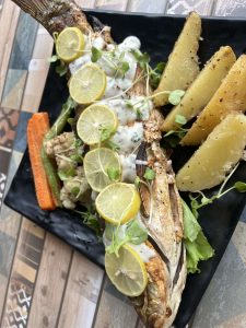 A beautifully presented whole grilled fish served on a black platter, draped in a creamy white sauce and topped with fresh lemon wheels and microgreens. Thick, seasoned potato wedges and grilled carrots line the side.