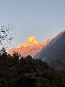 Sunlit mountain peak glowing with orange hues at sunrise, surrounded by darker forested slopes under a clear blue sky.