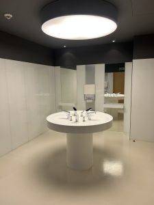 Modern and minimalist restroom interior featuring a circular white sink with multiple faucets, surrounded by mirrors and clean white surfaces. A large round ceiling light creates a bright, futuristic atmosphere. 
