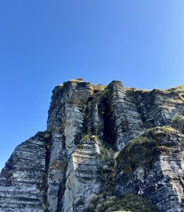 Layered gray rock cliffs rise dramatically against a deep blue cloudless sky, with textured stone, patches of grass, and moss emphasizing the rugged beauty and vertical composition of the landscape.
