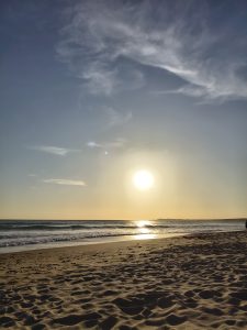 Sun setting over a tranquil beach, casting a warm glow on the sand. Gentle waves reflect the sunlight under a sky with wispy clouds. 
