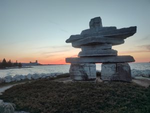 A large stone inukshuk structure stands prominently on a patch of low green shrubs along a shoreline. In the background, the sun has just set over a wide, wavy body of water, leaving a vibrant orange and red glow stretching across the horizon under a pale twilight sky. 