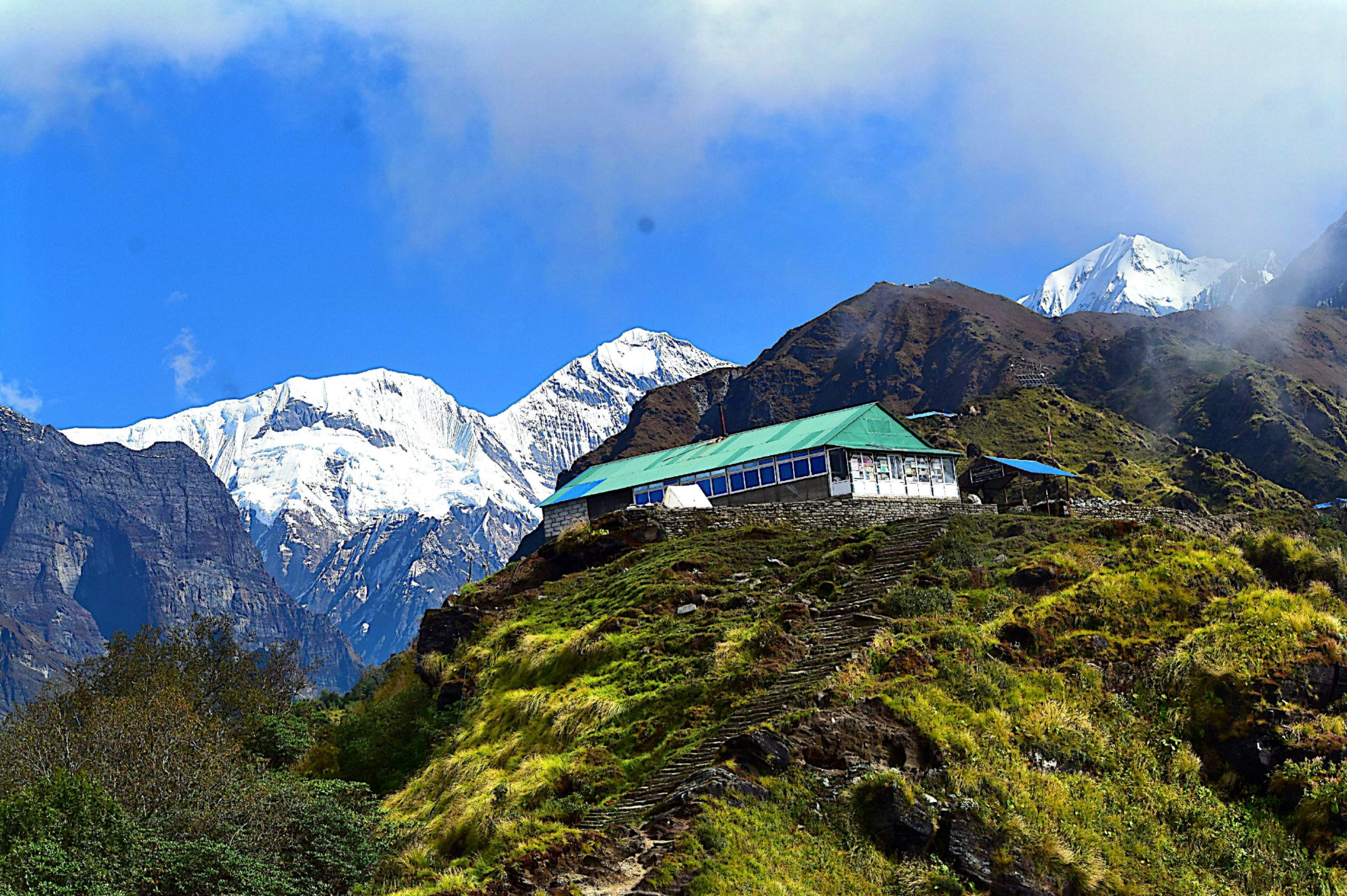 A small building with a green roof sits atop a lush hillside, set against towering snow-capped mountains under a bright blue sky with scattered clouds.