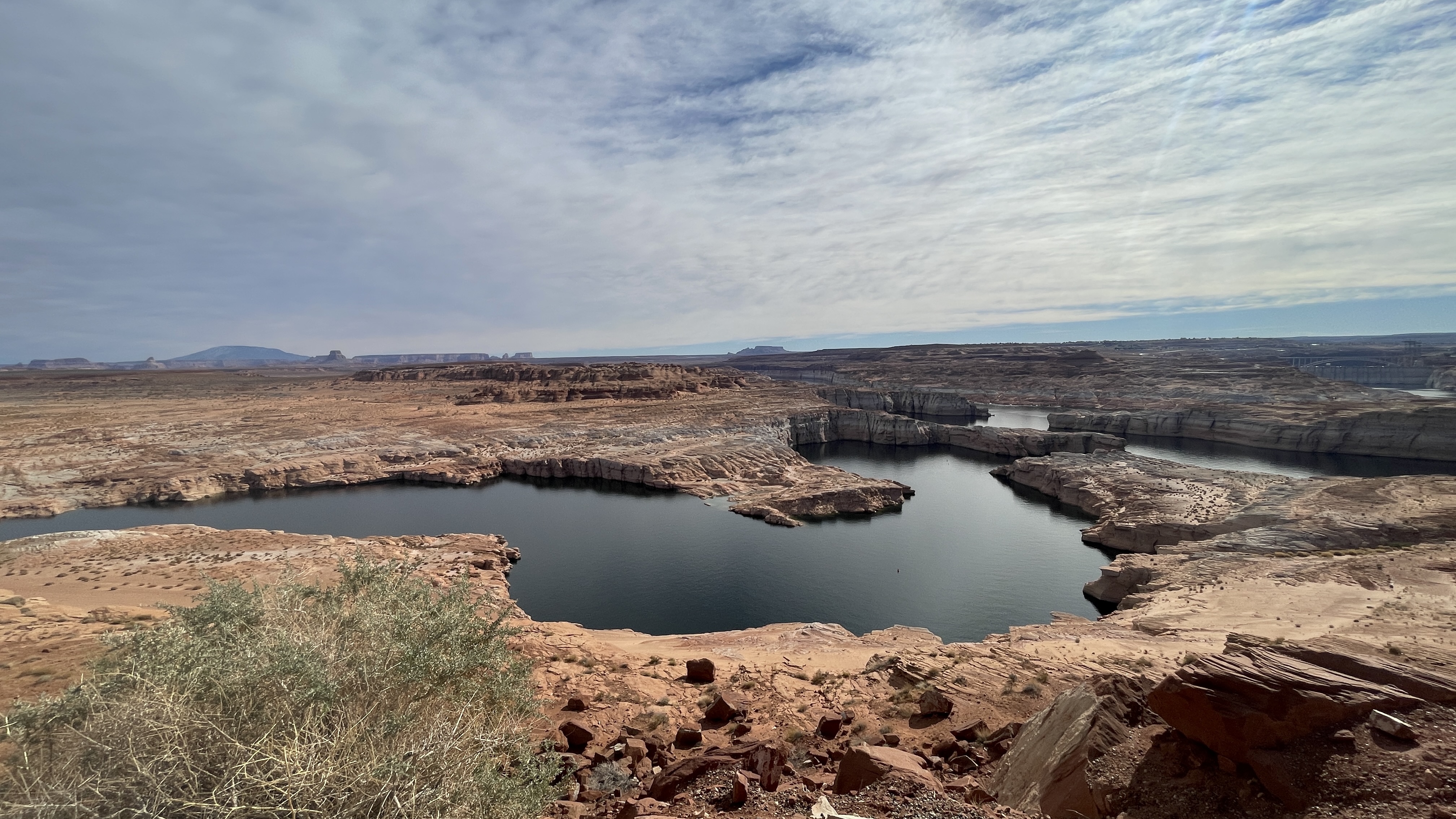 High-angle overview of a stark desert landscape featuring the dark blue waters of Lake Powell cutting through layers of orange and tan sandstone cliffs.