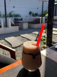 A frothy, blended coffee or chocolate frappe drizzled with chocolate syrup and served with a bold red straw sits on a wooden table. The background shows a bright, open-air café courtyard with gravel ground and comfortable white booth seating. 