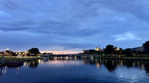 Evening river scene with a historic skyline reflected in calm water. A lit bridge arches overhead, its glow mirrored below. Trees line the bank beneath a deep blue, cloud-scattered twilight sky.
