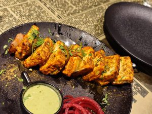 Grilled paneer tikka arranged on a black plate, garnished with herbs. Accompanied by a small bowl of green chutney and sliced red onions.