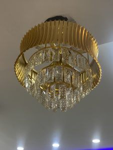 A circular, tiered gold chandelier with radiating metal fins and hanging crystal pendants, suspended from a white ceiling with recessed lights nearby.