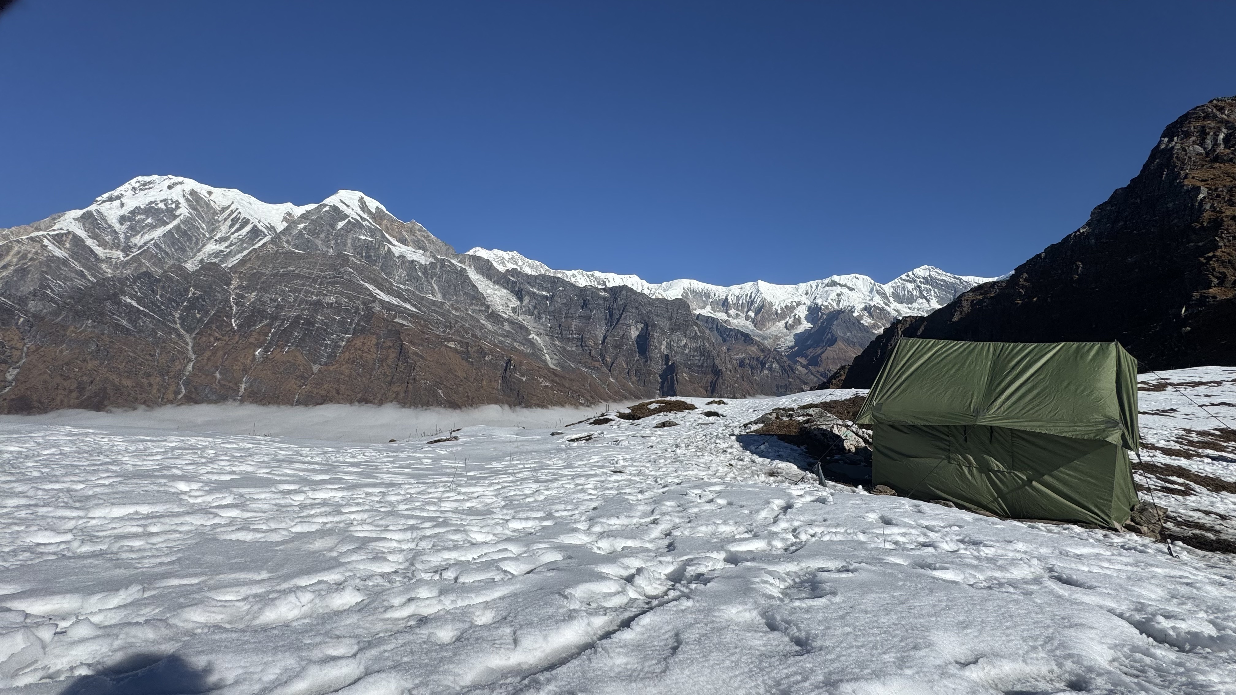 A bright daytime landscape of a snow-covered terrain leading toward a majestic range of rocky, snow-capped mountains under a clear blue sky. A plain green camping tent sits on the right side of the snowy foreground.