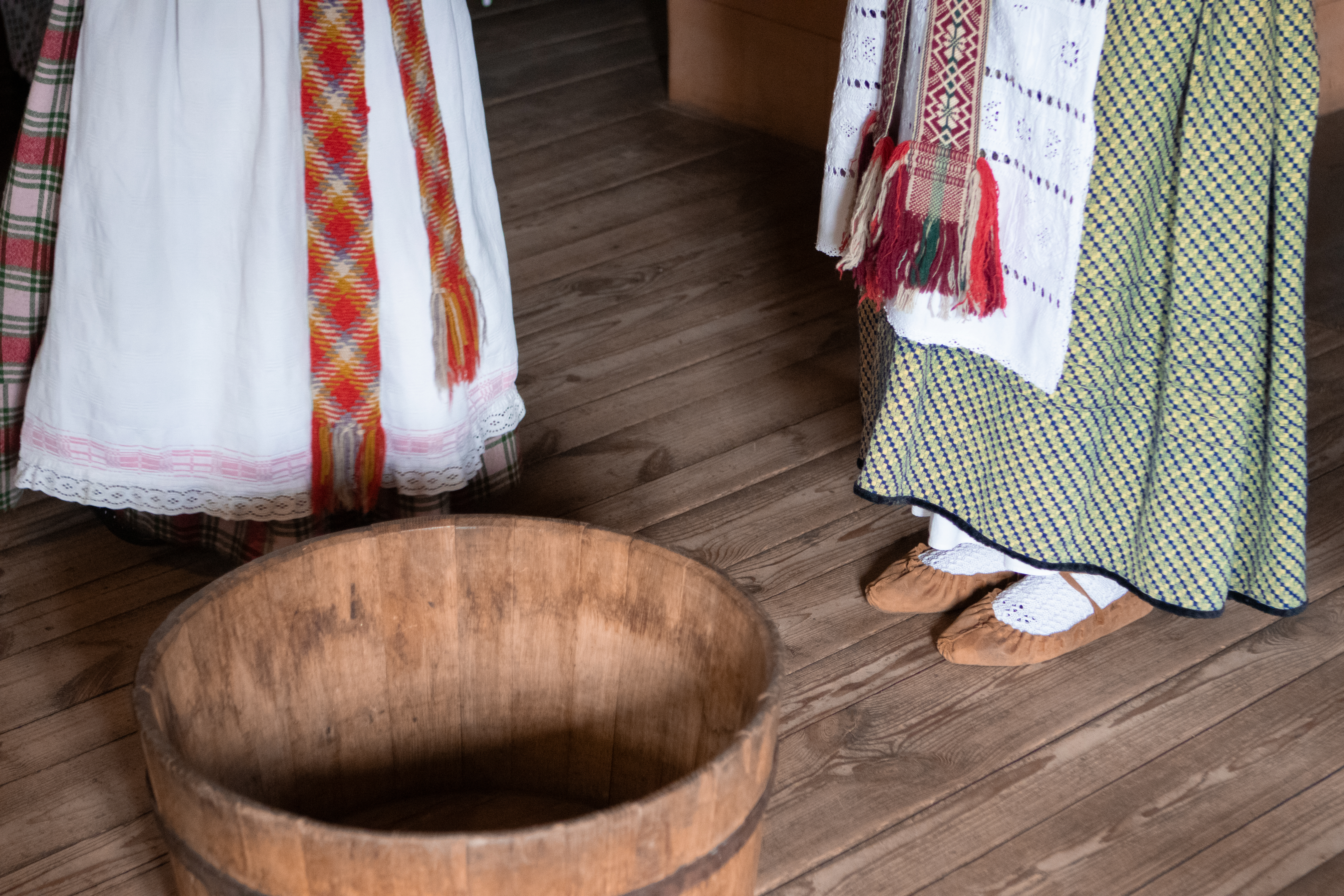The feet of the two women in traditional Lithuanian folk dresses standing beside a wooden pot for the bread making on a wooden floor in the wooden traditional farm house.
