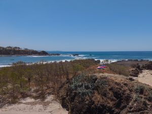 A Costa Rican flag stands on a rocky coastal point overlooking a blue ocean and small sandy beach. Dry vegetation, dark rocks, and white waves fill the foreground, with a clear blue sky above.