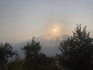 Sun setting behind clouds over a mountain landscape, casting a soft glow above silhouetted trees in the foreground