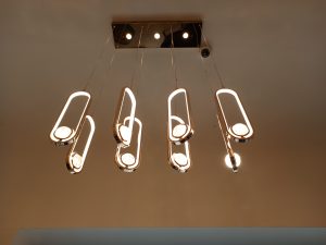 A contemporary indoor ceiling light fixture featuring a polished rectangular metallic base plate with four built-in spotlights and eight hanging, elongated oval rings lined with glowing LED strips that suspend textured glass discs.