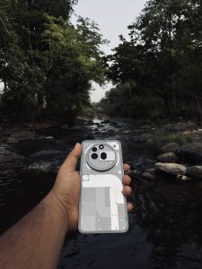 A hand holds a transparent smartphone, showing internal components, against a serene forest backdrop with a flowing stream.