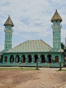 A beautiful mint-green mosque (Ayikkara Muhyidheen Masjid / Moideen Palli) featuring two matching minarets on either side. The central building and the minaret towers are topped with distinct, decorative, patterned roofs of yellow, green, and blue tiles. Captured at Kannur, Kerala.