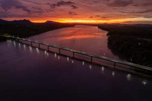An aerial view of a long bridge crossing a wide river at sunset. The sky glows in orange, purple, and pink tones, reflecting on the water, while dark mountains and forested land stretch into the distance.