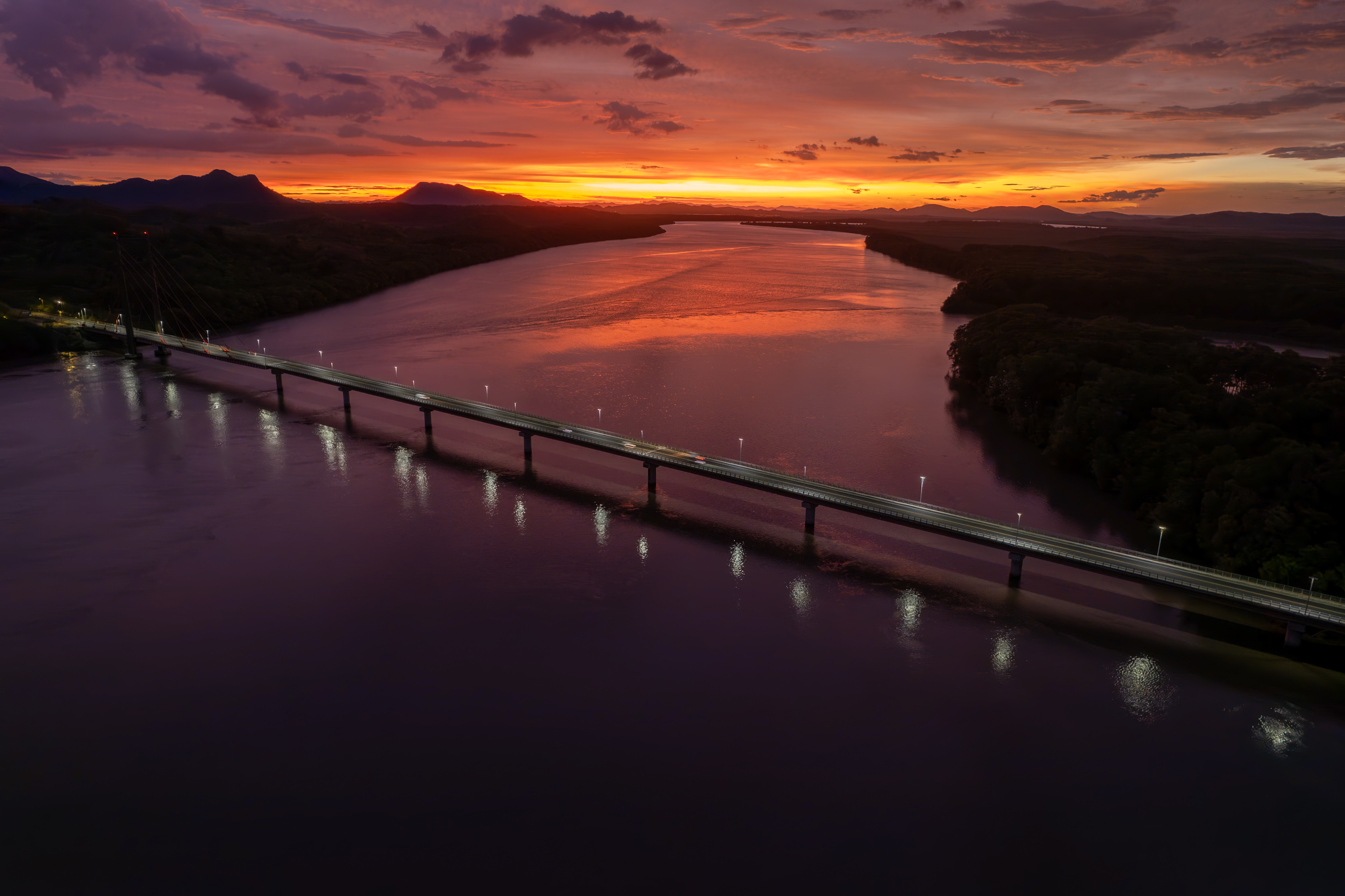An aerial view of a long bridge crossing a wide river at sunset. The sky glows in orange, purple, and pink tones, reflecting on the water, while dark mountains and forested land stretch into the distance.