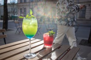 Two colorful iced drinks with mint garnish on a sunny outdoor café table, with a blurred person and city street in the background.
