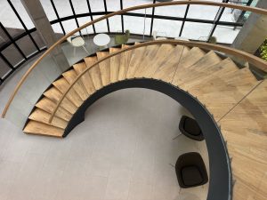 Elegant spiral staircase with wooden steps and glass railing descends to a modern seating area with two chairs and a small table below.