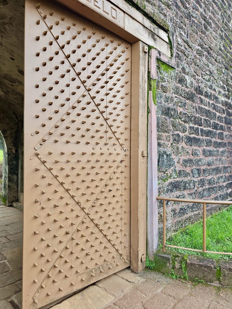 A heavy metal-studded iron door stands beside thick laterite walls, showcasing the strong defensive design of the historic fort. Captured at St. Angelo Fort, Kannur, Kerala.