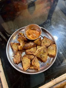 Spiced fried fokso (yam/taro) wedges served on a stainless steel plate with a small cup of tomato-chili dipping sauce.